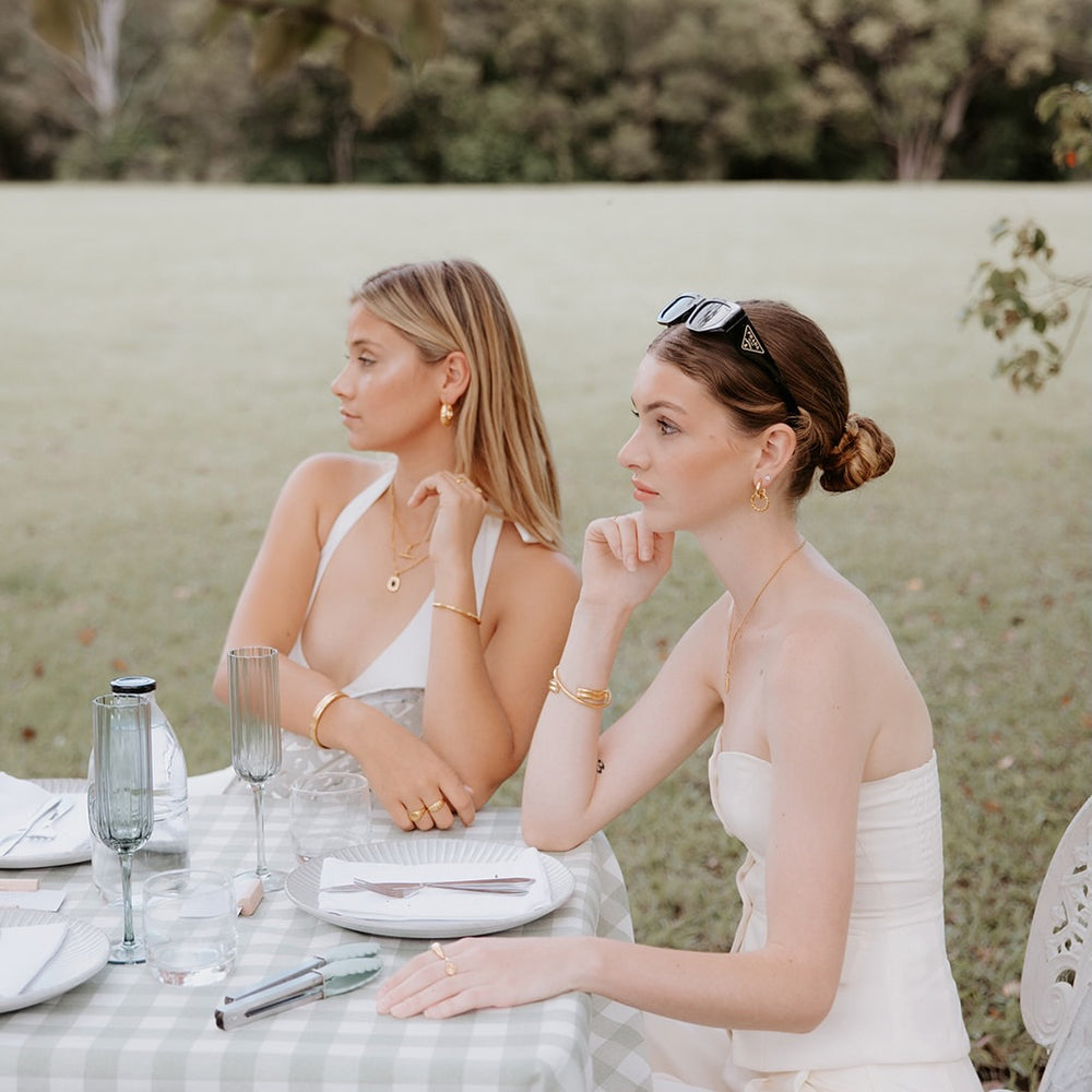Two women sitting at a table outdoors, wearing white dresses and waterproof gold jewellery. designer sunglasses
