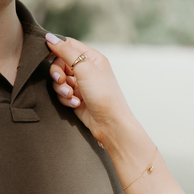 Hand with a gold ring and bracelet adjusting a brown shirt collar.